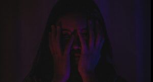 Asian woman in a darkly lit room with her hands over her face