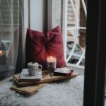 reading nook by the window with a mug, a red pillow, a lit candle, and a fuzzy seating pillow